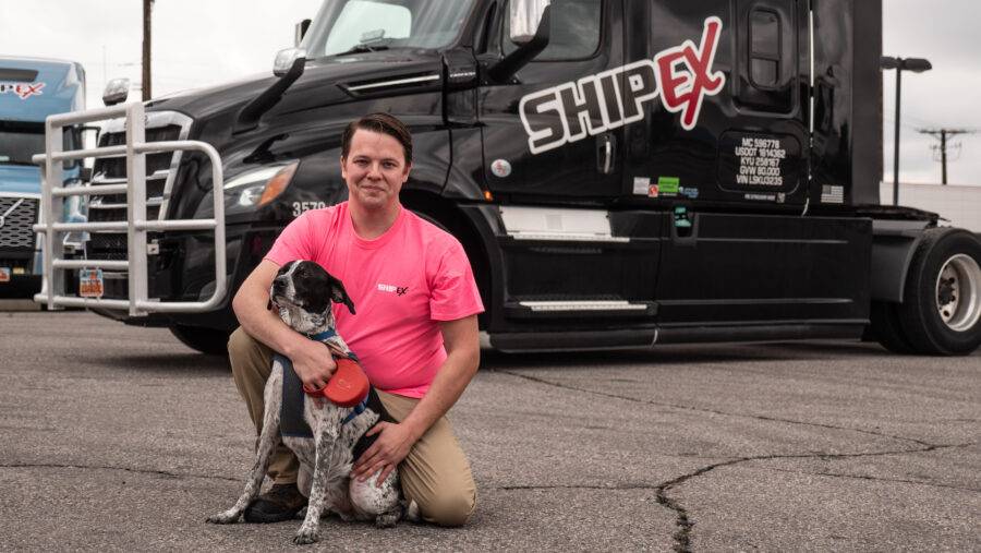 truck driver wearing a pink shirt kneeling in front of a black semi truck with dog