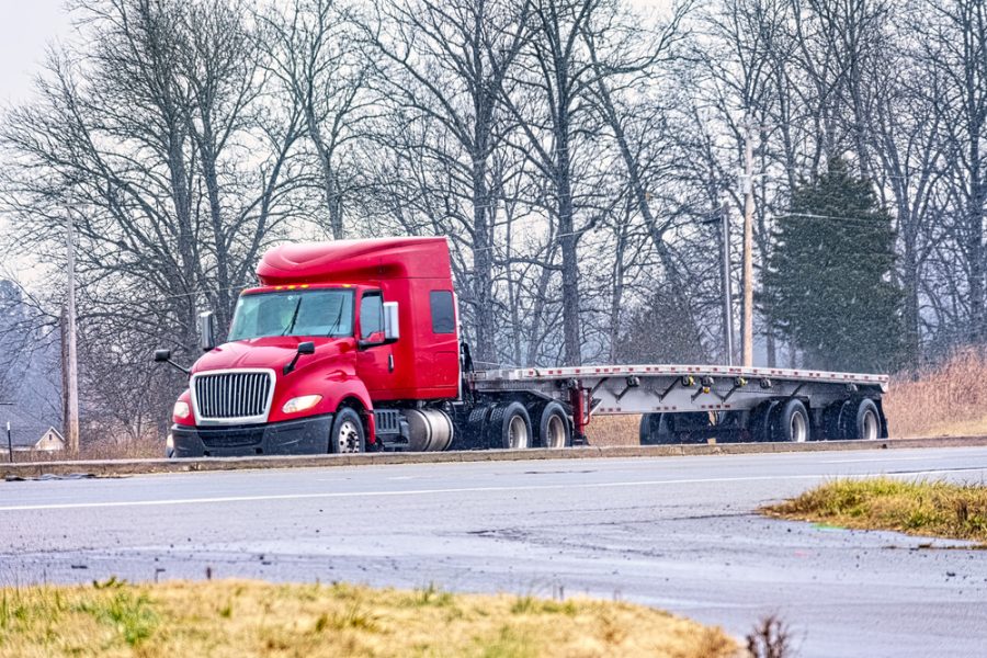 Horizontal shot of an empty flatbed tractor trailer driving in mixed winter weather.